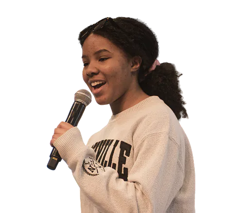 Young woman with curly hair tied back, wearing a sweater and glasses on her head, speaking into a microphone with a smile.